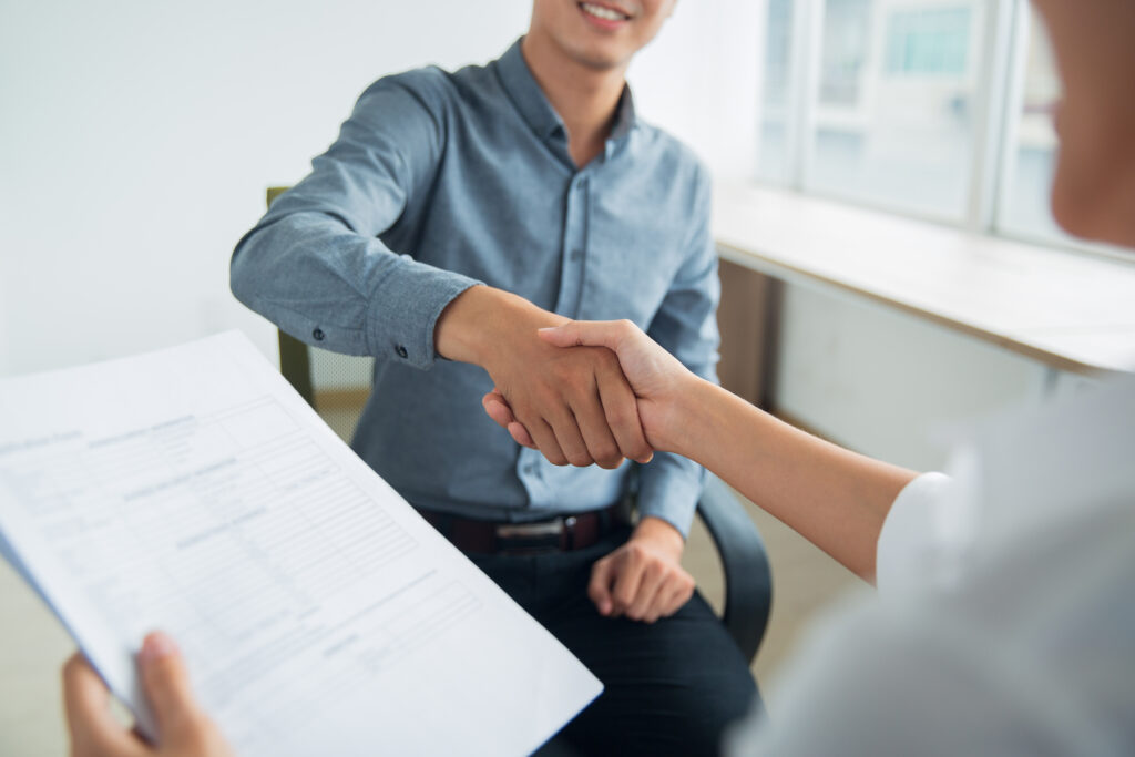 Smiling Asian businessman wearing shirt sitting in office and shaking hand of female partner. Woman holding document